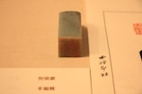 A rectangular stone stamp with an intricate red carving sits on a beige surface. To its side, there are two sheets of paper with text written in a script that appears to be East Asian characters.