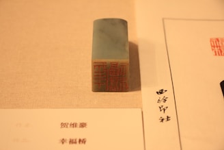 A rectangular stone stamp with an intricate red carving sits on a beige surface. To its side, there are two sheets of paper with text written in a script that appears to be East Asian characters.