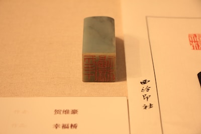 A rectangular stone stamp with an intricate red carving sits on a beige surface. To its side, there are two sheets of paper with text written in a script that appears to be East Asian characters.