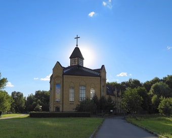 A large, historic-looking church building with a prominent cross on top of a steep roof. The architecture features tall, arched windows and intricate brickwork. The building is surrounded by lush greenery and a well-kept lawn, under a bright, clear blue sky with a few clouds.