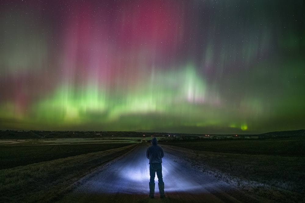 a man standing in the middle of a road under an aurora display