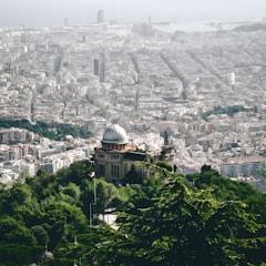 A sprawling cityscape view of Barcelona, featuring a mix of historic and modern architecture. In the foreground, an observatory with a domed roof is surrounded by lush green trees. The city stretches out toward the sea, with distinct grid patterns formed by its streets.