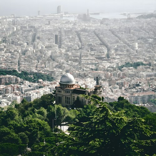A sprawling cityscape view of Barcelona, featuring a mix of historic and modern architecture. In the foreground, an observatory with a domed roof is surrounded by lush green trees. The city stretches out toward the sea, with distinct grid patterns formed by its streets.