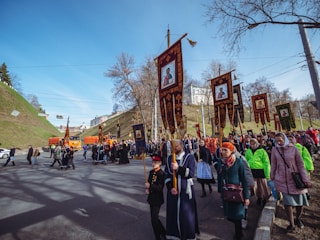 A large group of people are participating in a religious procession, marching along a street. They are holding ornate banners depicting religious icons. Many participants are dressed in traditional or religious clothing, including robes and headscarves. The scene is set in a city area with grassy hills and trees in the background, under a clear blue sky.