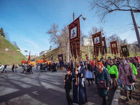 A large group of people are participating in a religious procession, marching along a street. They are holding ornate banners depicting religious icons. Many participants are dressed in traditional or religious clothing, including robes and headscarves. The scene is set in a city area with grassy hills and trees in the background, under a clear blue sky.