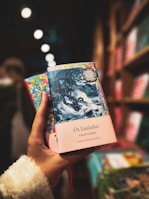 A hand is holding a colorful book titled 'Os Lusiadas' by Luis de Camões. The book cover features an intricate design, with a blend of dark blues and light hues. The background shows a blurred bookshelf filled with other books, creating a cozy and inviting atmosphere.