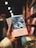 A hand is holding a colorful book titled 'Os Lusiadas' by Luis de Camões. The book cover features an intricate design, with a blend of dark blues and light hues. The background shows a blurred bookshelf filled with other books, creating a cozy and inviting atmosphere.