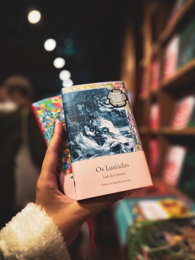 A hand is holding a colorful book titled 'Os Lusiadas' by Luis de Camões. The book cover features an intricate design, with a blend of dark blues and light hues. The background shows a blurred bookshelf filled with other books, creating a cozy and inviting atmosphere.