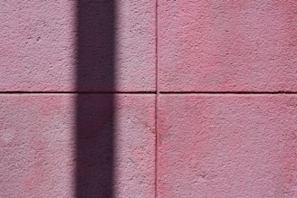 A textured pink wall composed of large square tiles, with a prominent vertical shadow cast across its surface, adding contrast and depth.