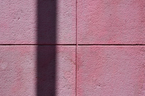 A textured pink wall composed of large square tiles, with a prominent vertical shadow cast across its surface, adding contrast and depth.