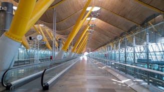 A spacious and modern airport terminal features a series of large, yellow structural beams that support a high, curved ceiling made of wood paneling. Glass walls with metallic frameworks run along the sides, allowing natural light to illuminate the interior. A moving walkway extends down the center, guiding travelers along the bright and airy corridor. A large clock and several lighting fixtures hang from the ceiling, contributing to the functionality of the space.