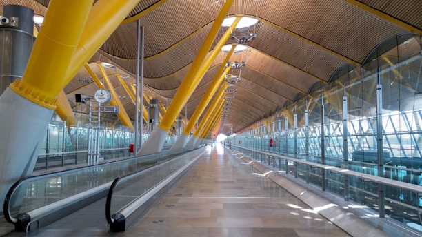 A spacious and modern airport terminal features a series of large, yellow structural beams that support a high, curved ceiling made of wood paneling. Glass walls with metallic frameworks run along the sides, allowing natural light to illuminate the interior. A moving walkway extends down the center, guiding travelers along the bright and airy corridor. A large clock and several lighting fixtures hang from the ceiling, contributing to the functionality of the space.