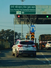A white SUV decorated with ribbons is stopped in front of a toll booth that has signs indicating slow-moving and toll-exempt vehicles. The road also has other vehicles including a truck covered with a tarp and another white car. Traffic lights and various road signs are visible.
