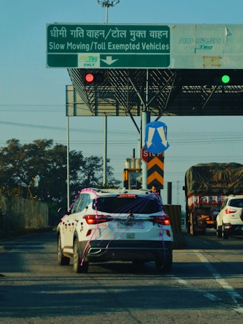 A white SUV decorated with ribbons is stopped in front of a toll booth that has signs indicating slow-moving and toll-exempt vehicles. The road also has other vehicles including a truck covered with a tarp and another white car. Traffic lights and various road signs are visible.