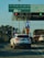A white SUV decorated with ribbons is stopped in front of a toll booth that has signs indicating slow-moving and toll-exempt vehicles. The road also has other vehicles including a truck covered with a tarp and another white car. Traffic lights and various road signs are visible.