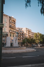 A roundabout with a stone monument featuring human figures is surrounded by buildings with balconies and shutters. Trees line the street, and vehicles are parked around the area. The sky is clear, and the scene is well-lit by daylight.