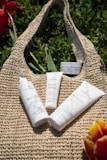 Several skincare products are placed on a woven bag surrounded by green foliage. The containers have white packaging with black and green text, and there are bursts of color from red and yellow flowers at the edges.