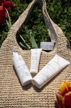 Several skincare products are placed on a woven bag surrounded by green foliage. The containers have white packaging with black and green text, and there are bursts of color from red and yellow flowers at the edges.