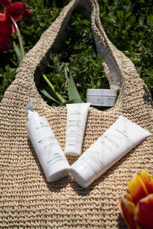 Several skincare products are placed on a woven bag surrounded by green foliage. The containers have white packaging with black and green text, and there are bursts of color from red and yellow flowers at the edges.