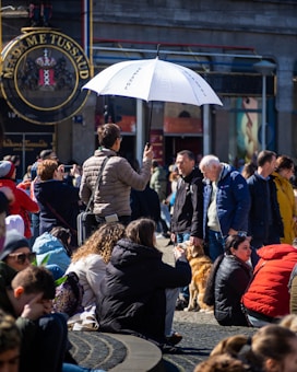 A bustling outdoor scene with people gathered around, some sitting and others standing. A person with a white umbrella that has text printed on it is prominently visible, suggesting they may be guiding a tour group. The background features the signage for Madame Tussaud's, indicating a popular tourist location. People are dressed in winter or early spring attire, and a dog is present among the crowd.