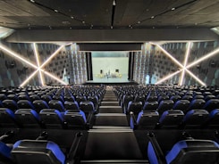 A modern theater with rows of blue and black seats facing a stage. The walls have geometric patterns with illuminated lines creating an 'X' shape. The stage is empty except for a few people standing near the back.