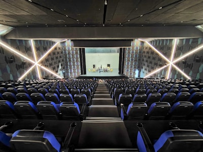 A modern theater with rows of blue and black seats facing a stage. The walls have geometric patterns with illuminated lines creating an 'X' shape. The stage is empty except for a few people standing near the back.