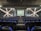 A modern theater with rows of blue and black seats facing a stage. The walls have geometric patterns with illuminated lines creating an 'X' shape. The stage is empty except for a few people standing near the back.