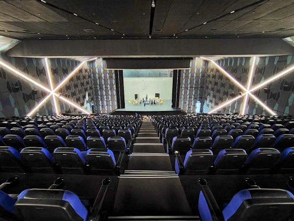 A modern theater with rows of blue and black seats facing a stage. The walls have geometric patterns with illuminated lines creating an 'X' shape. The stage is empty except for a few people standing near the back.