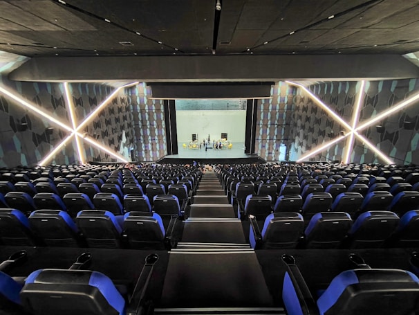 A modern theater with rows of blue and black seats facing a stage. The walls have geometric patterns with illuminated lines creating an 'X' shape. The stage is empty except for a few people standing near the back.