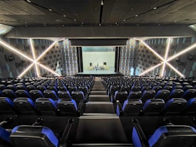 A modern theater with rows of blue and black seats facing a stage. The walls have geometric patterns with illuminated lines creating an 'X' shape. The stage is empty except for a few people standing near the back.