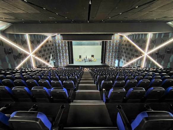A modern theater with rows of blue and black seats facing a stage. The walls have geometric patterns with illuminated lines creating an 'X' shape. The stage is empty except for a few people standing near the back.