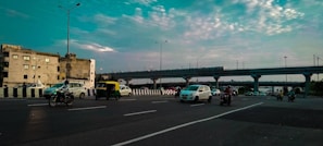 A busy urban road with several vehicles, including cars, motorcycles, and an auto-rickshaw, moving in both directions. A train is visible on an elevated track in the background, while buildings and a partly cloudy sky complete the scene.