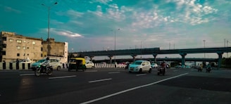 A busy urban road with several vehicles, including cars, motorcycles, and an auto-rickshaw, moving in both directions. A train is visible on an elevated track in the background, while buildings and a partly cloudy sky complete the scene.