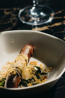 A dish features a grilled sausage topped with crumbled cheese and served on a bed of greens. The image has a dark, moody ambiance with a soft focus glass of water or wine in the background. The lighting creates a play of shadows and highlights on the dish, enhancing its textures.