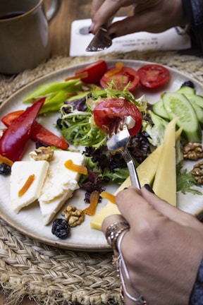 A hand holds a piece of tomato with a fork above a plate filled with vibrant vegetables, cheeses, and nuts. The plate includes slices of cucumber, tomato, red and green peppers, mixed greens, walnuts, and chunks of cheese. Dried fruits are scattered around the plate, all placed on a woven mat.
