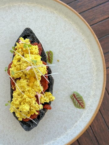 A black tortilla filled with bright yellow scrambled topping and red sauce, garnished with sprouts and microgreens, placed on a textured beige plate. The backdrop features a wooden texture.
