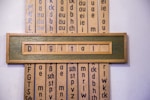 A wooden framed artwork displaying the word 'Digital' in blue letters set against a muted green background. Vertical panels in the background contain various combinations of letters and phonetic symbols in blue on a beige surface.