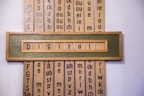 A wooden framed artwork displaying the word 'Digital' in blue letters set against a muted green background. Vertical panels in the background contain various combinations of letters and phonetic symbols in blue on a beige surface.