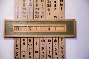 A wooden framed artwork displaying the word 'Digital' in blue letters set against a muted green background. Vertical panels in the background contain various combinations of letters and phonetic symbols in blue on a beige surface.