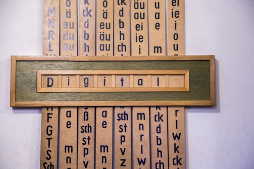 A wooden framed artwork displaying the word 'Digital' in blue letters set against a muted green background. Vertical panels in the background contain various combinations of letters and phonetic symbols in blue on a beige surface.
