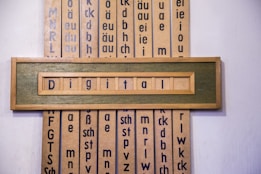 A wooden framed artwork displaying the word 'Digital' in blue letters set against a muted green background. Vertical panels in the background contain various combinations of letters and phonetic symbols in blue on a beige surface.