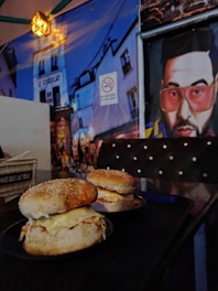 A dimly lit restaurant scene featuring two burgers with sesame seed buns and melted cheese on a small table. The background includes a vibrant wall mural depicting a street scene and a stylized portrait of a person wearing sunglasses. A sign on the wall indicates no smoking.