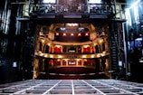 An elaborate theater interior viewed from the stage with ornate balconies and red seats. The theater is dimly lit with a few bright lights illuminating the balconies. The stage area appears to be under maintenance with visible metal framework.
