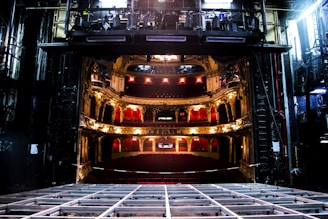 An elaborate theater interior viewed from the stage with ornate balconies and red seats. The theater is dimly lit with a few bright lights illuminating the balconies. The stage area appears to be under maintenance with visible metal framework.