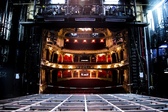 An elaborate theater interior viewed from the stage with ornate balconies and red seats. The theater is dimly lit with a few bright lights illuminating the balconies. The stage area appears to be under maintenance with visible metal framework.
