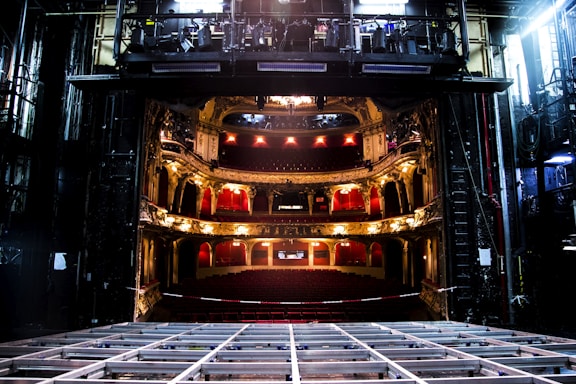An elaborate theater interior viewed from the stage with ornate balconies and red seats. The theater is dimly lit with a few bright lights illuminating the balconies. The stage area appears to be under maintenance with visible metal framework.