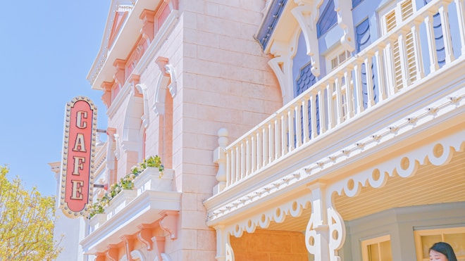A building with ornate architectural details features a sign labeled 'CAFE' in large, bold letters. The structure has a blend of pastel colors, primarily light pink and blue, accented with white trim and decorative elements. Greenery is present on the balcony, adding a touch of nature to the scene. The overall aesthetic is reminiscent of a vintage or whimsical style.