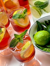 A collection of beverage glasses filled with a vibrant red drink, adorned with lemon slices and fresh mint leaves. The drinks are served in ribbed glassware, and there is a bowl containing whole limes and more mint leaves to the side.