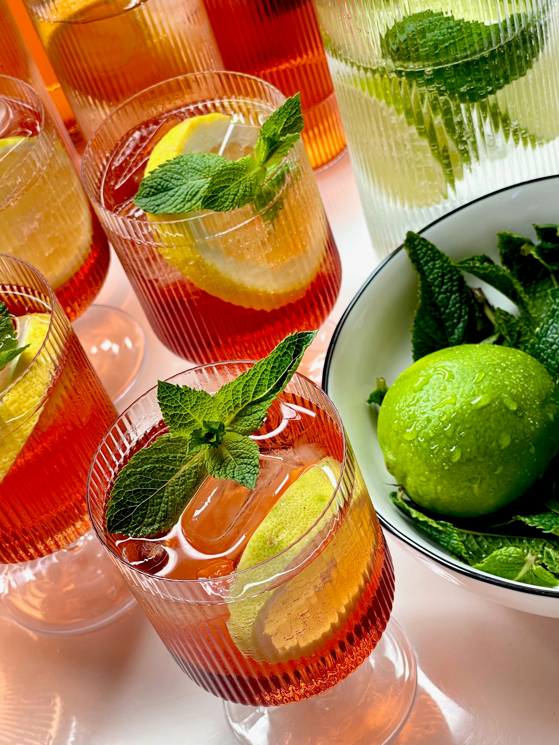 A collection of beverage glasses filled with a vibrant red drink, adorned with lemon slices and fresh mint leaves. The drinks are served in ribbed glassware, and there is a bowl containing whole limes and more mint leaves to the side.