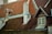 The image features a series of steeply pitched roofs with red clay tiles and wooden accents. There is a prominent white chimney, flanked by weathered roofing. A window with detailed wood framing is visible, hinting at a rustic architectural style.